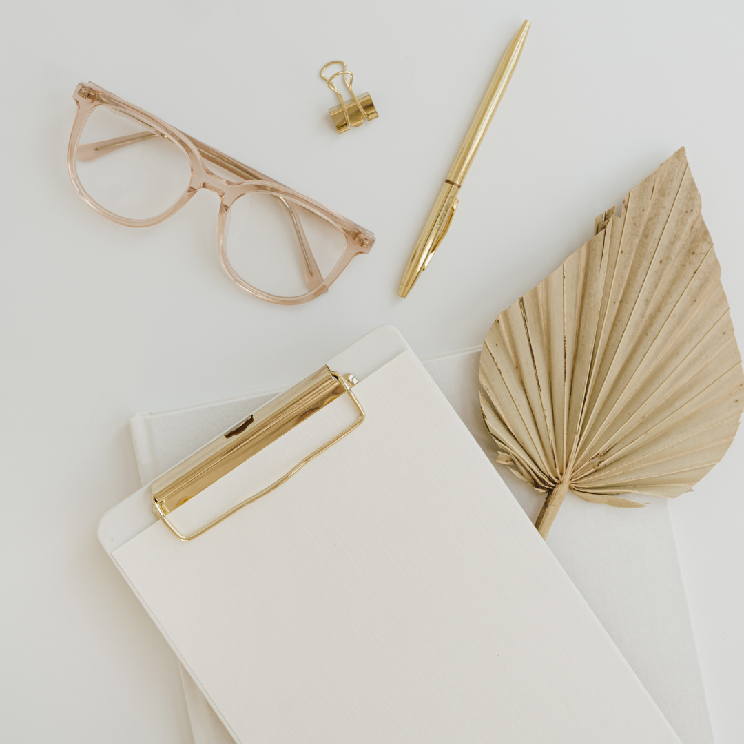 Flat lay of a neutral-toned workspace with clipboard, gold pen, eyeglasses, and a dried palm leaf—representing calm, minimalist content strategy and aligned creative flow.