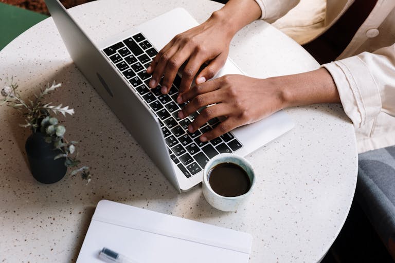 Close-up of a woman’s hands typing on a laptop at a round white table with a cup of coffee nearby—capturing a calm, focused moment of content creation and digital work.