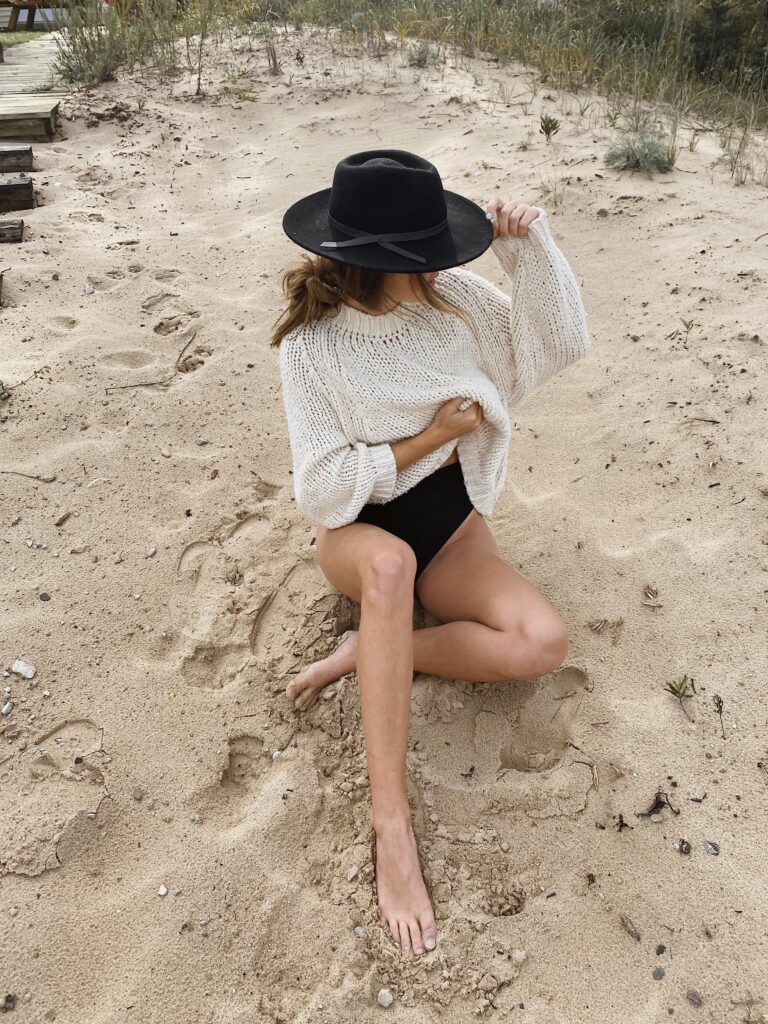 Pregnant Mom sitting on sandy beach wearing a loose knit sweater, black swimsuit, and wide-brimmed black hat, with her face partially hidden as she holds the brim. Natural grasses and wooden steps visible in the background.