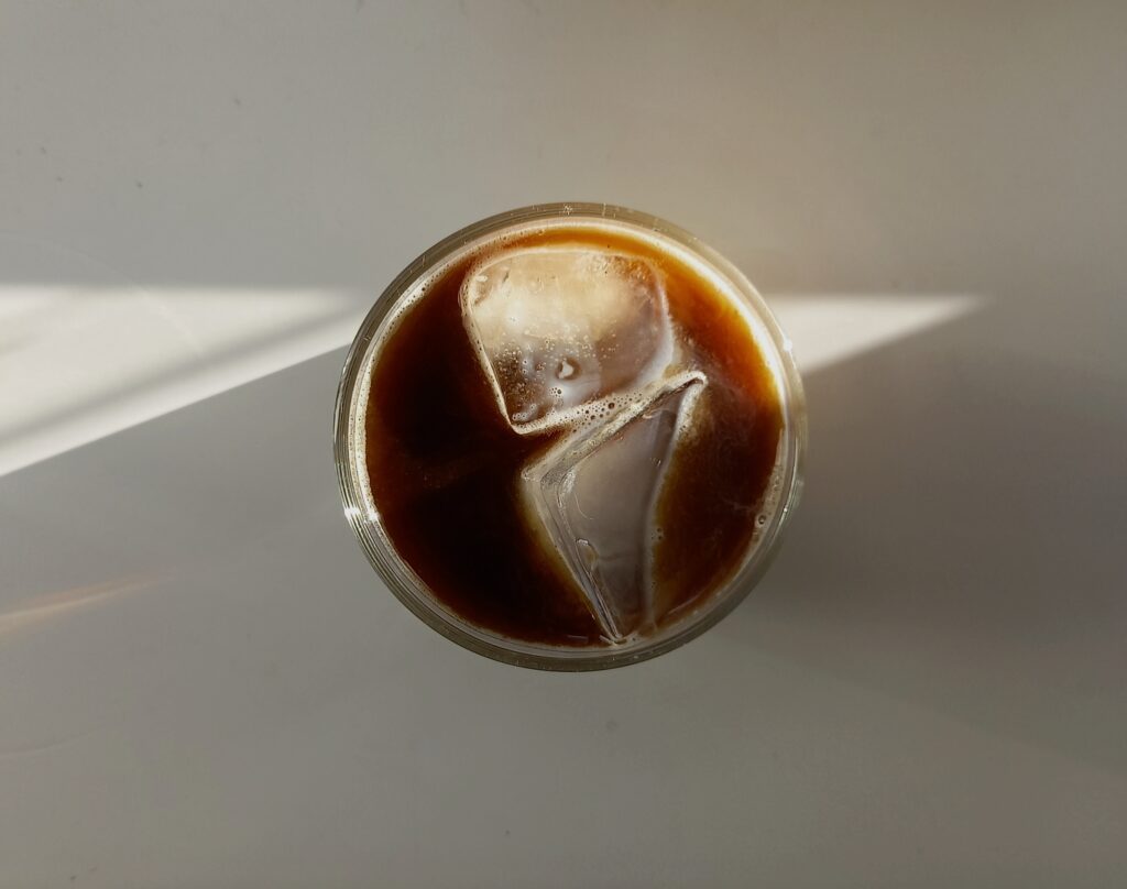 Top-down view of a glass of iced black coffee with two large ice cubes, resting on a light surface with soft natural sunlight casting diagonal shadows.
