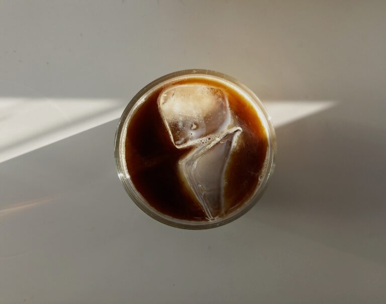 Top-down view of a glass of iced black coffee with two large ice cubes, resting on a light surface with soft natural sunlight casting diagonal shadows.