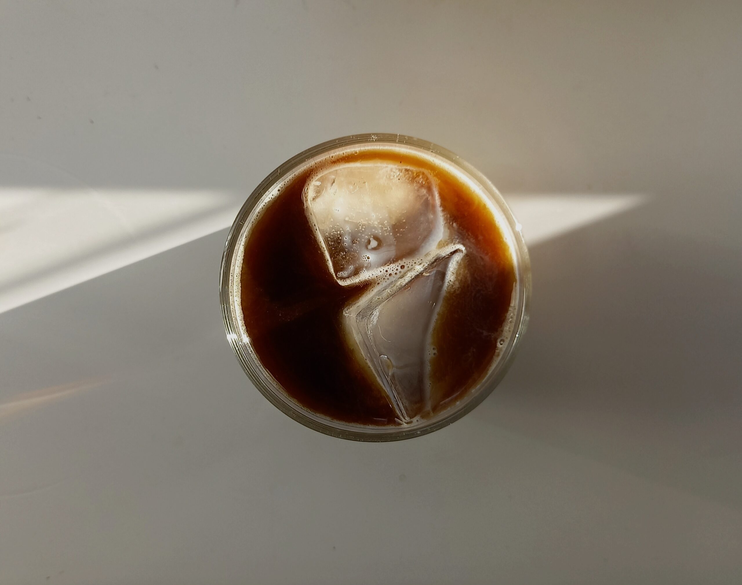 Top-down view of a glass of iced black coffee with two large ice cubes, resting on a light surface with soft natural sunlight casting diagonal shadows.